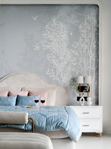 Bed with blue linens and pink pillows against a grey tree-mural wall, white nightstand with lamps beside it