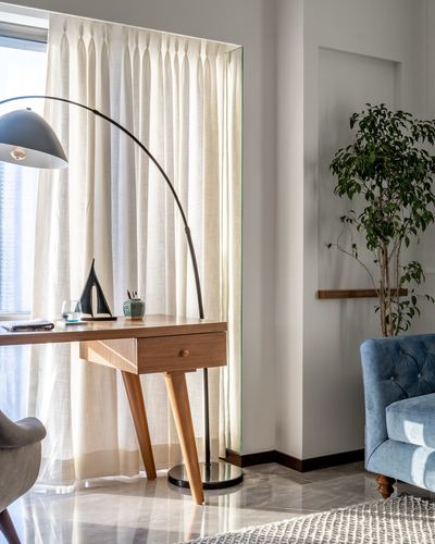 Wooden desk with arc lamp beside sheer curtains and a blue tufted armchair on marble floor
