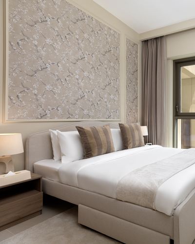 Beige upholstered bed against floral wallpaper panel with striped pillows and bedside lamp