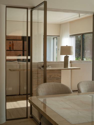Glass pivot door opens onto beige living room with stone console and lamp, Dubai apartment