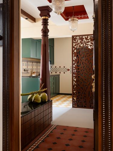 View through open carved wooden door past a turned pillar into a green kitchen with tiled floors