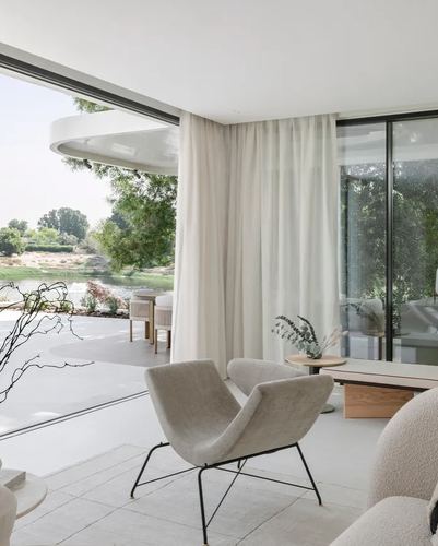 Minimalist living room with sheer curtains, curved armchair, and glass wall opening to a garden and lake