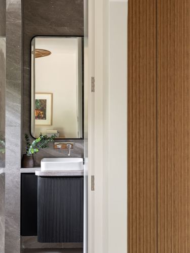 Doorway view of a stone-clad powder room with black fluted vanity and mirror reflecting living space