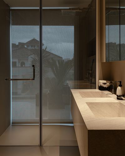 Bathroom vanity with stone counter and glass shower wall overlooking villas at dusk in Dubai