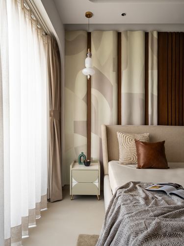 Bedroom corner with beige bed, leather and knit pillows, wooden pendant lamp, and sheer curtains