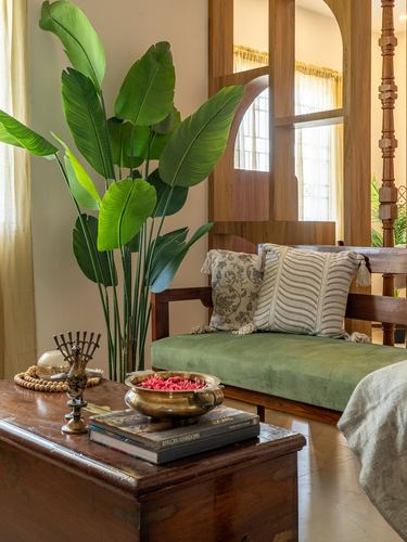 Green velvet bench with patterned cushions beside carved wooden trunk and large plant