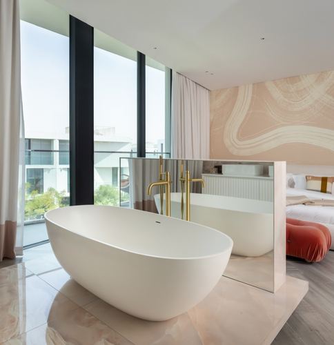 Freestanding oval tub with gold floor faucet before floor-to-ceiling windows, mirror partition to bedroom beyond