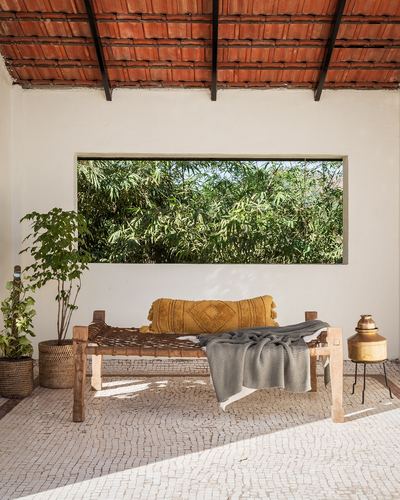 Woven wooden daybed with mustard cushion under a long window framed by greenery, terracotta tiled roof above