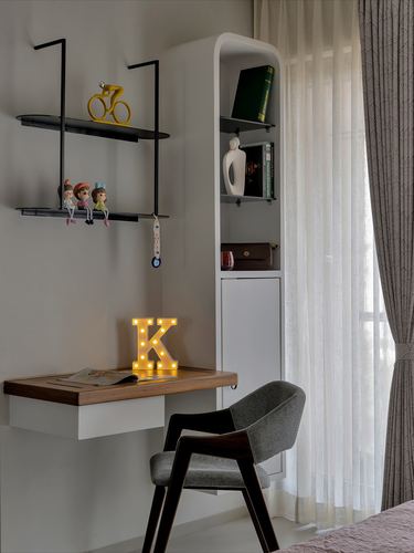 Floating wood desk with marquee letter light beneath black wall shelves near curved white cabinet and sheer curtains