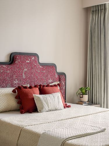Bed with patterned maroon headboard, tasseled cushions, and sage curtains beside a nightstand with a plant