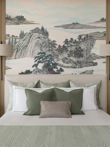 Bed with sage and white pillows beneath a painted mountain landscape mural, flanked by wall sconces