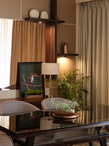 Dining table vignette with framed bird art, lamp, and plant against curtained window and wood partition.