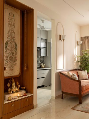 Wooden pooja niche with painted panel and brass idol beside living room with sofa