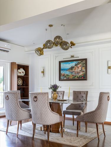 Dining room with marble table, quilted taupe chairs, brass chandelier and framed coastal painting on panelled wall