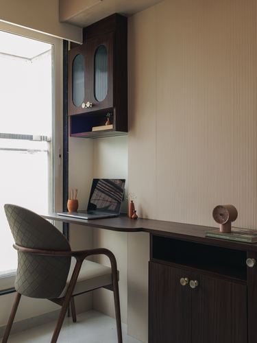 Built-in wooden desk under window with woven chair and overhead fluted-glass cabinet