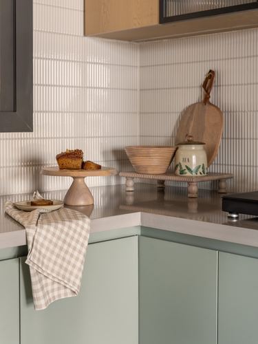 Kitchen counter corner with wooden cake stand, tea canister and ribbed tile backsplash above sage cabinets