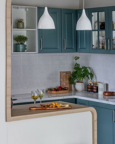 Teal kitchen cabinetry with white pendant lights and curved wood breakfast counter styled with plants and snacks