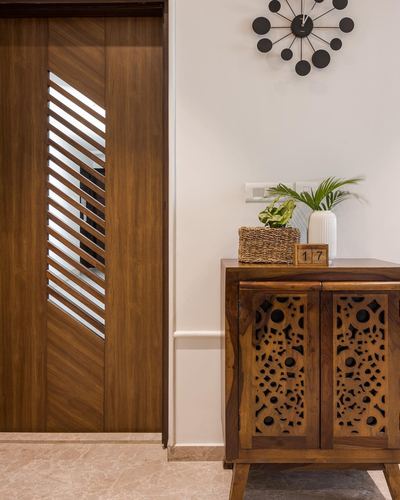 Wooden louvered door beside carved-panel cabinet with clock and plants in foyer