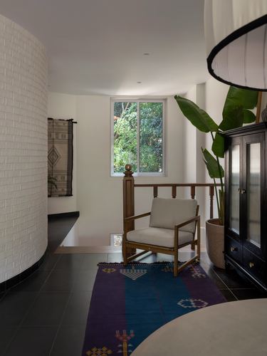 Curved white brick landing with wooden armchair, kilim rug, black cabinet and window view of greenery.