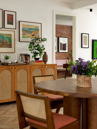 Dining area with cane sideboard, gallery wall, round wood table and brick doorway beyond