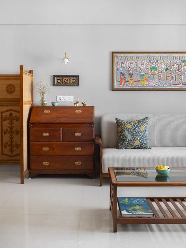 Living room with vintage wooden bureau, cane screen, sofa, and framed madhubani painting on grey wall