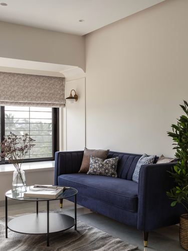 Navy velvet sofa against cream wall beside arched window with patterned roman blind and glass coffee table