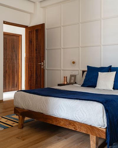 Bedroom with panelled white wall, wooden bed with navy linens, and open teak door to hallway