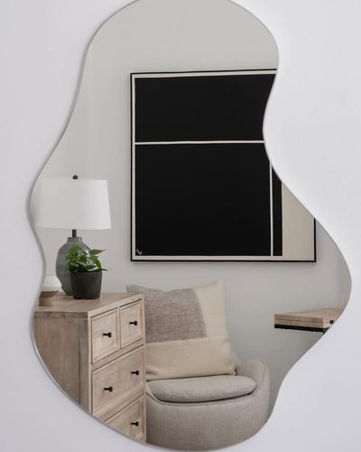 Organic-shaped mirror reflecting a beige armchair, wood dresser, lamp, and black abstract art
