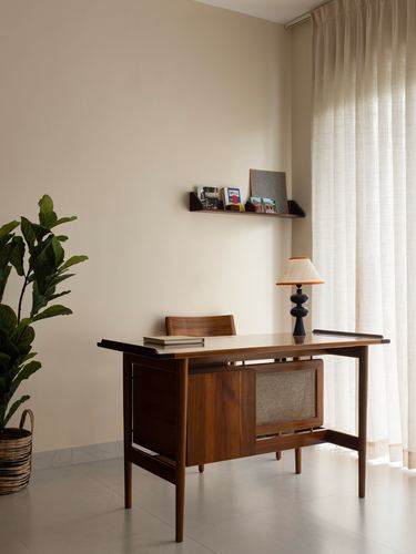 Mid-century wooden desk with lamp and shelf against beige wall near sheer curtain
