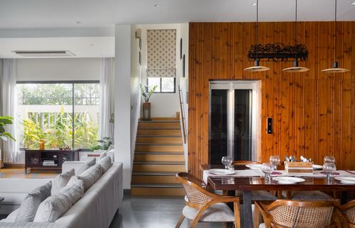 Open-plan living and dining space with wood-panelled wall, staircase, and glass elevator in Raipur home