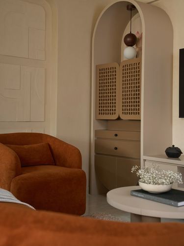 Arched niche with perforated cabinet doors beside a terracotta velvet armchair in a lime-plastered living room
