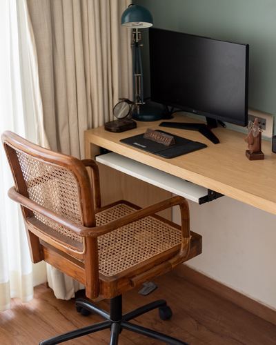Cane armchair beside wall-mounted wooden desk with monitor and lamp against sage wall
