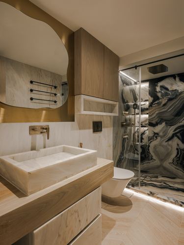 Bathroom with stone vessel sink, wood cabinetry, curved brass mirror, and dark veined stone shower wall