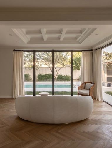 Round cream sofa in living room with herringbone wood floor facing glass doors to pool and garden