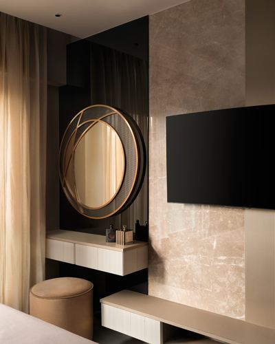 Round brass-framed mirror over fluted dressing table beside marble-clad TV wall in bedroom