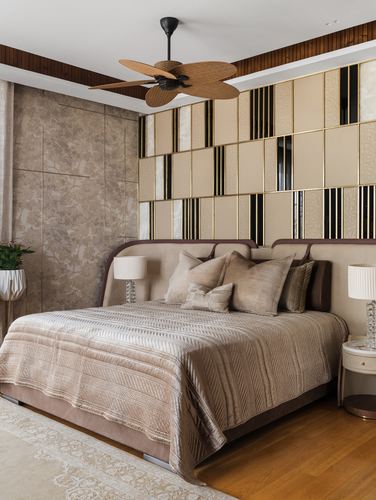 Beige bedroom with upholstered bed, patterned gold wall panel, and rattan ceiling fan