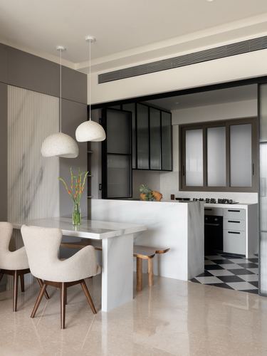 Dining area with marble table, boucle chairs, dome pendants, and glimpse of checkerboard-floor kitchen beyond glass partition