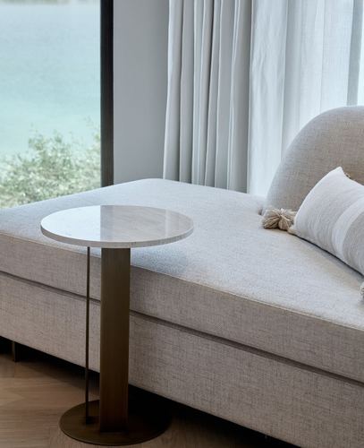 Round stone side table on brass base beside a beige upholstered daybed near a window with a water view
