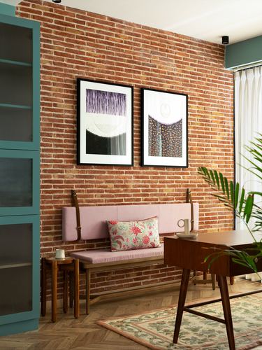 Exposed brick wall with framed art above a pink upholstered bench and wooden desk in foreground