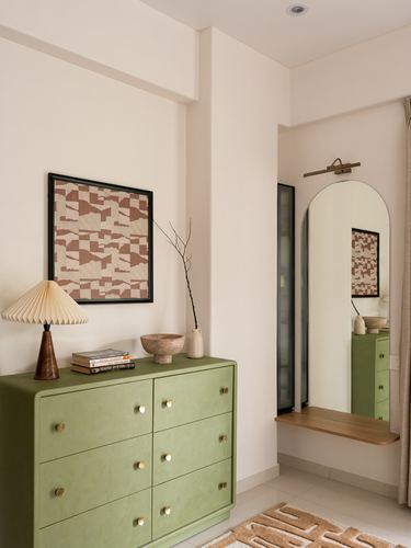 Sage green six-drawer dresser with brass knobs below a framed patterned textile, beside an arched mirror niche