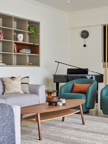 Living room with grey sofa, teal armchairs, oval wood coffee table and a black grand piano against beige walls