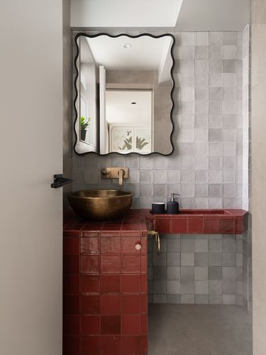 Powder room with brass vessel sink on red tiled vanity, wavy mirror, grey tiled wall