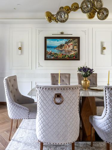 Dining room with quilted grey velvet chairs, marble table, and framed coastal painting on panelled wall.