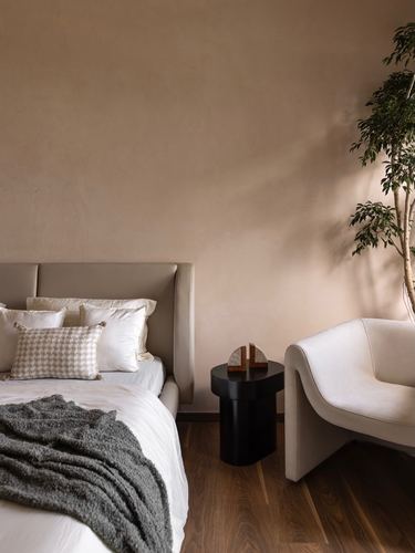 Warm-toned bedroom corner with upholstered bed, dark wood floor, curved armchair and potted plant.