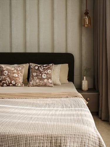 Bed with dark upholstered headboard, floral cushions, waffle throw, and brass pendant beside curtained window.