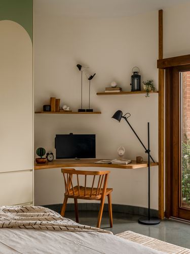 Corner study nook with curved wooden desk, floating shelves, spindle chair, and arched window