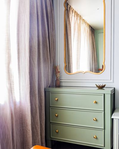 Sage green wooden dresser with brass knobs beneath a scalloped mirror, sheer curtains beside window