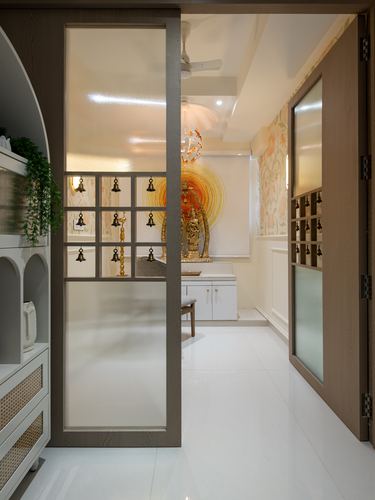 Frosted glass sliding door with brass bells opens to a backlit pooja alcove with glowing mural
