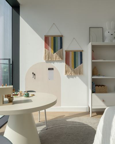 Child's bedroom corner with striped fringed wall hangings, arched niche, white bookshelf and play table