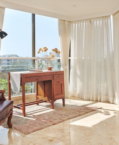 Wooden writing desk with dried flowers by floor-to-ceiling window overlooking balcony, sheer curtains, marble floor